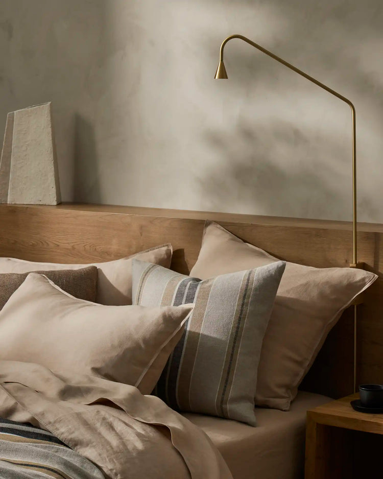 Neutral bedroom with Ravello Flat Sheet in shell, layered with striped linen pillows and taupe bedding, warm wooden bed frame, brass arc lamp, minimalist Scandinavian aesthetic