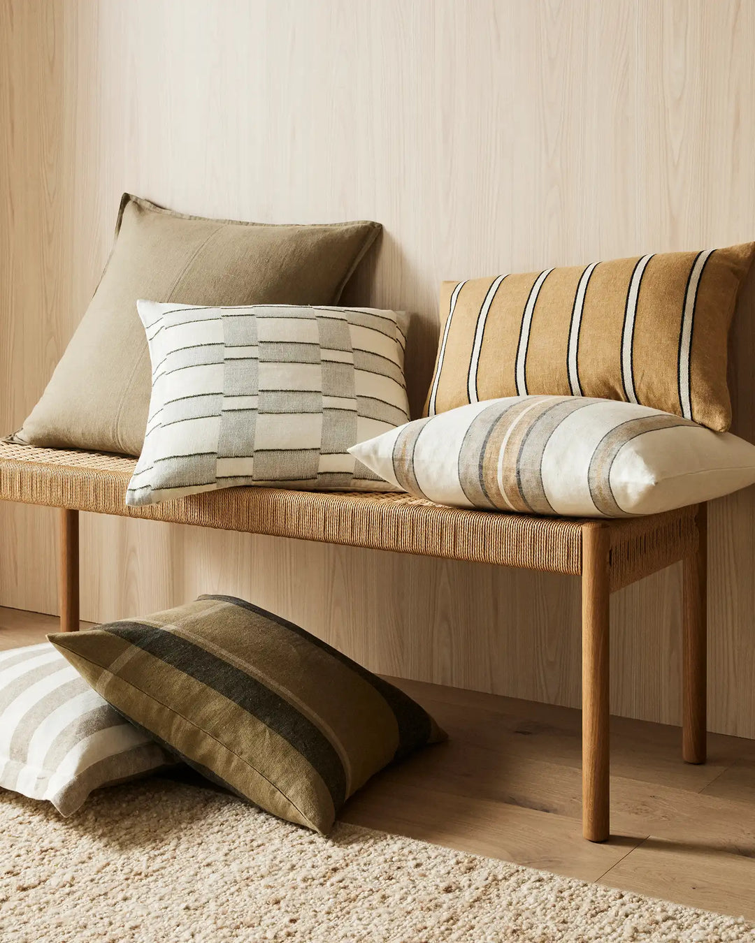 Warm-toned cushion arrangement on wooden bench: solid and striped linen pillows in cream, gold, and olive against light wood panelling and natural fibre rug