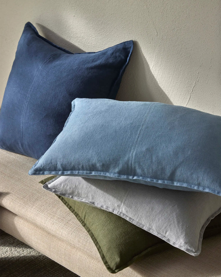 Layered linen cushions in navy, soft blue, stone and olive tones stacked against neutral textured wall, showcasing Como range in relaxed, monochromatic styling