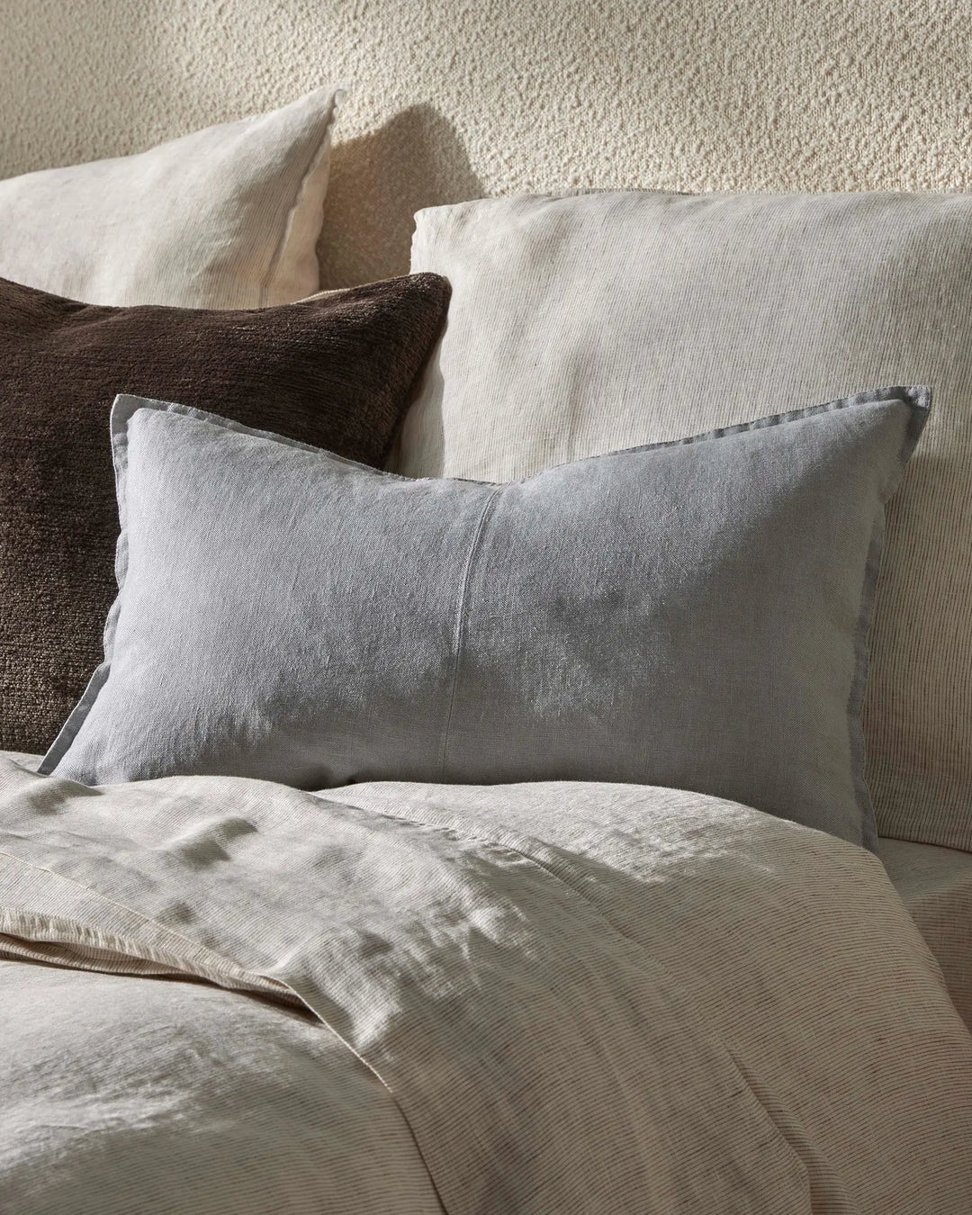 Neutral bedroom with layered pillows in cream, grey and charcoal tones, featuring textured linen and ribbed fabrics against a woven wall backdrop