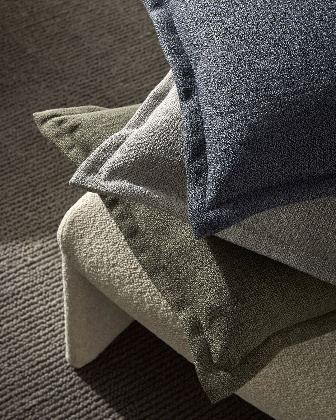 Neutral toned cushions in charcoal, sage and cream stacked on light wood chair against textured carpet, showcasing layered earth palette and boucle fabric texture