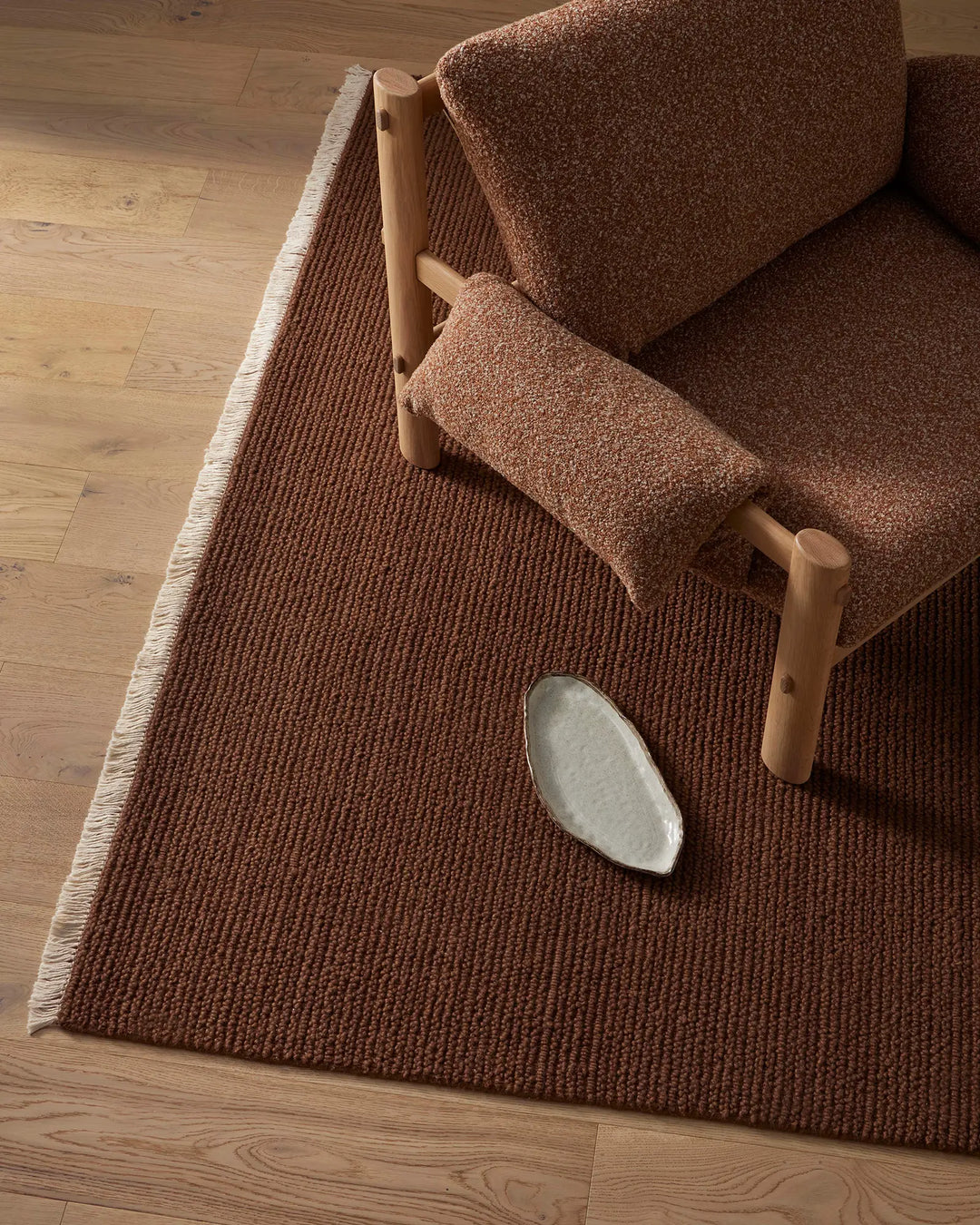 Warm terracotta wool rug with ribbed texture anchoring a minimalist living space, natural wood chair with linen upholstery and ceramic vessel styling