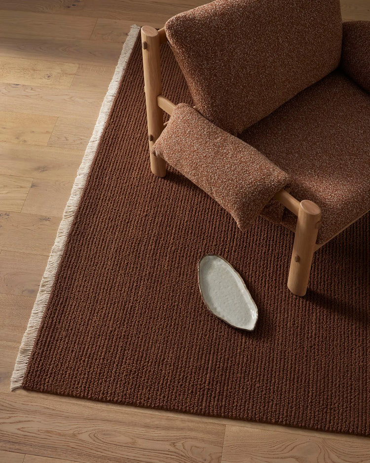 Warm terracotta wool rug with ribbed texture anchoring a minimalist living space, natural wood chair with linen upholstery and ceramic vessel styling