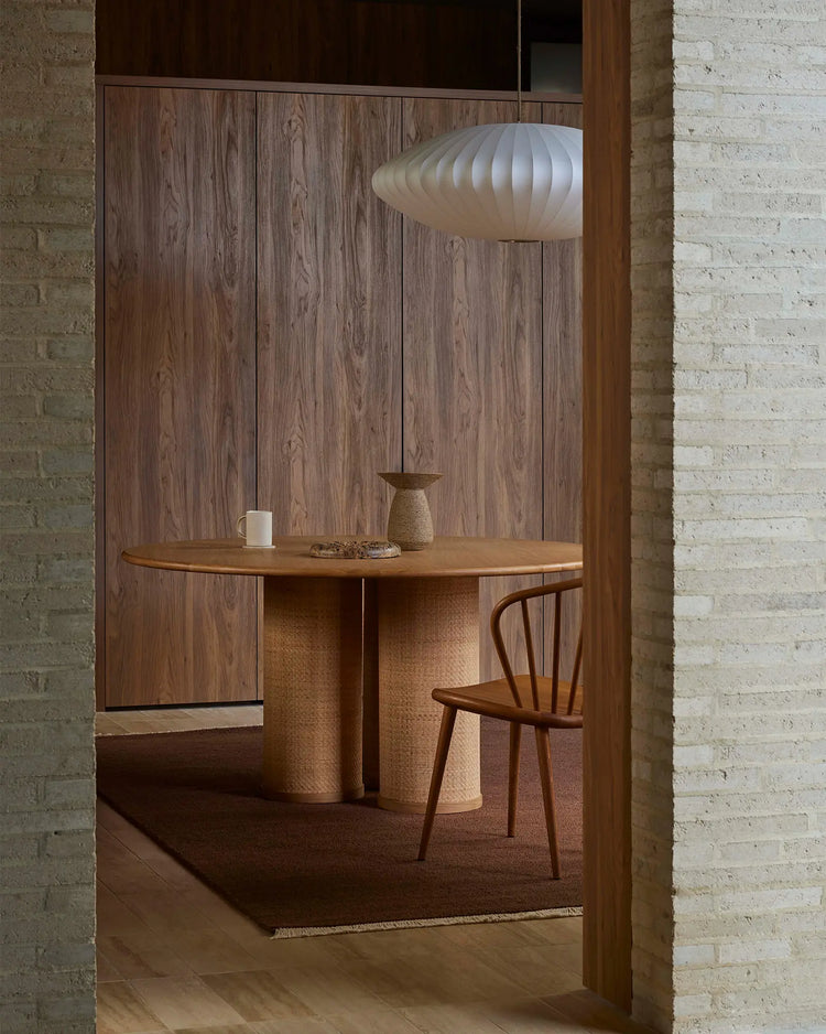 Warm timber dining nook with round wooden table, woven base, and mid-century chair beneath sculptural pendant, styled on terracotta wool rug against exposed brick backdrop