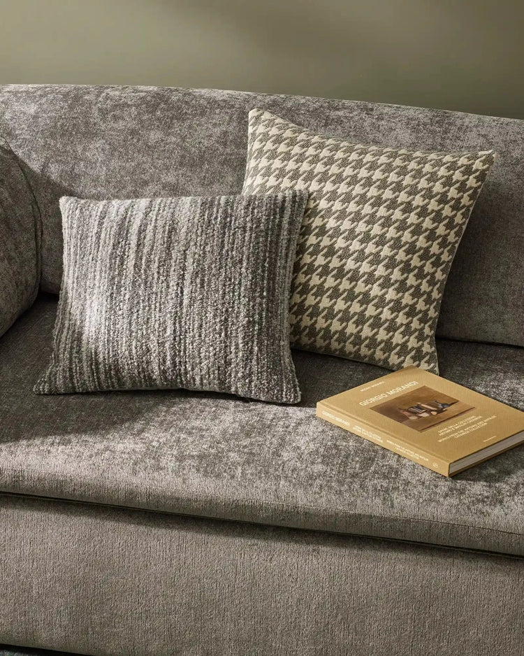 Neutral grey linen sofa styled with textured wool cushions in houndstooth and ribbed weave patterns, sage green wall backdrop, lifestyle scene with design book
