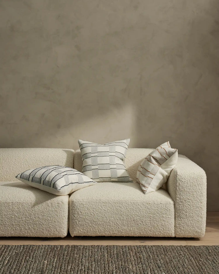 Minimalist living room styled with cream modular sofa, geometric stripe cushions in neutral and terracotta tones, textured jute rug, and soft natural light