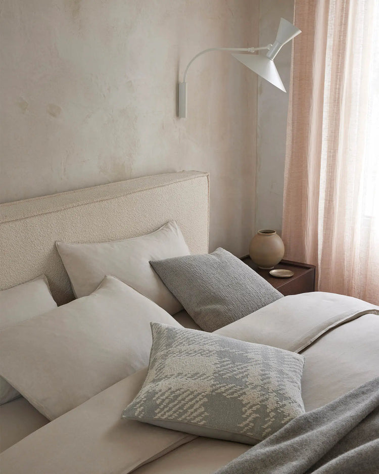 Serene neutral bedroom with cream linen headboard, layered white and grey textiles, patterned Laurent Smoke cushion, and soft pink curtains filtering natural light