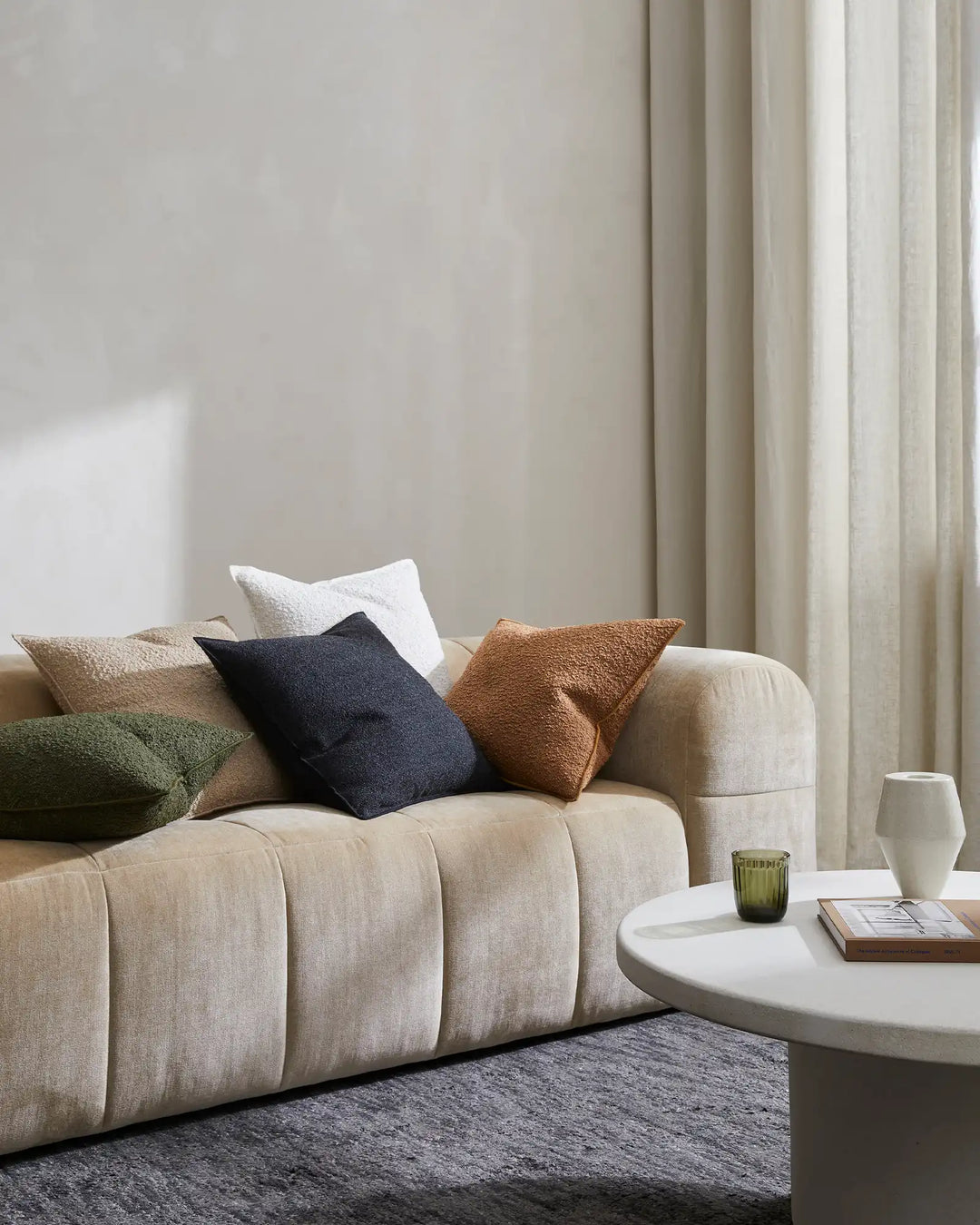 Neutral living room with layered Alberto cushions in ivory, midnight, copper and olive tones arranged on a beige sofa, Almonte Coal rug, floor-to-ceiling linen curtains, and minimalist styling