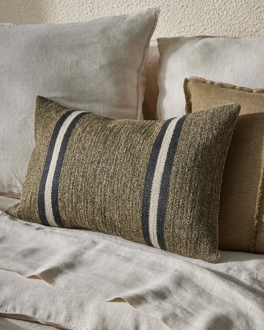 Neutral bedroom styling with Mila Forest cushion featuring woven texture and navy stripes, layered with cream linen pillows against a soft textured wall backdrop