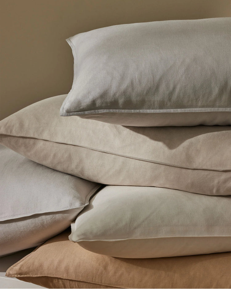 Stacked linen pillowcases in neutral tones—bone, biscuit, sage, shell and silver—showcase soft textural layering and earthy palette styling for minimalist bedding design.