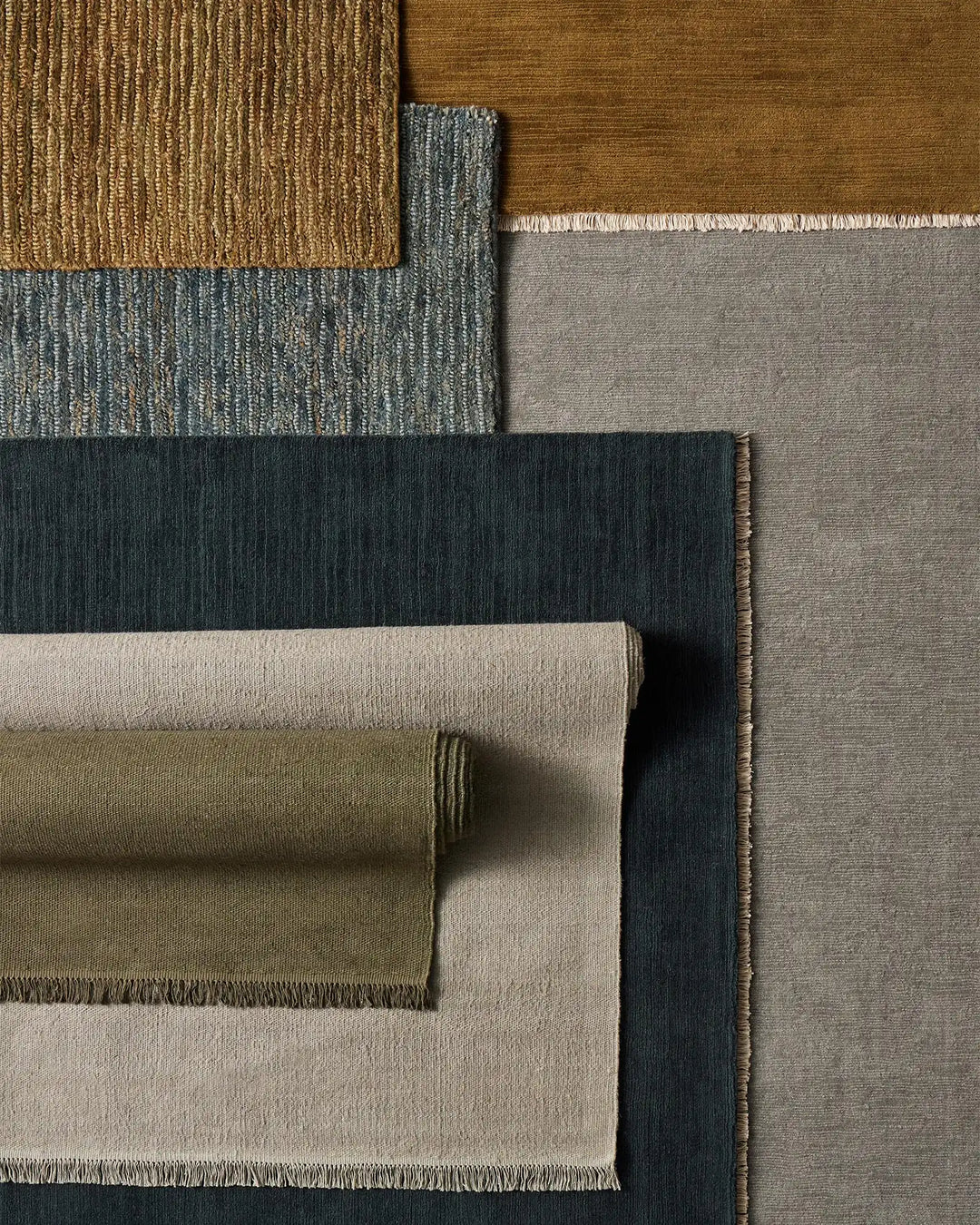 Layered neutral and earth-tone rugs in jute and wool showcase textural variety, from warm ochre and sage to cool charcoal and soft grey with woven and flat weave finishes