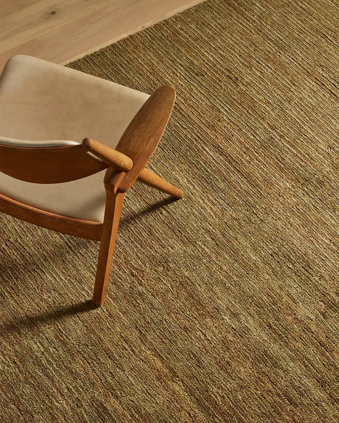 Mid-century wooden chair with cream upholstery on textured Bastian oakmoss jute rug, warm neutral palette with natural light casting shadows across woven floor covering