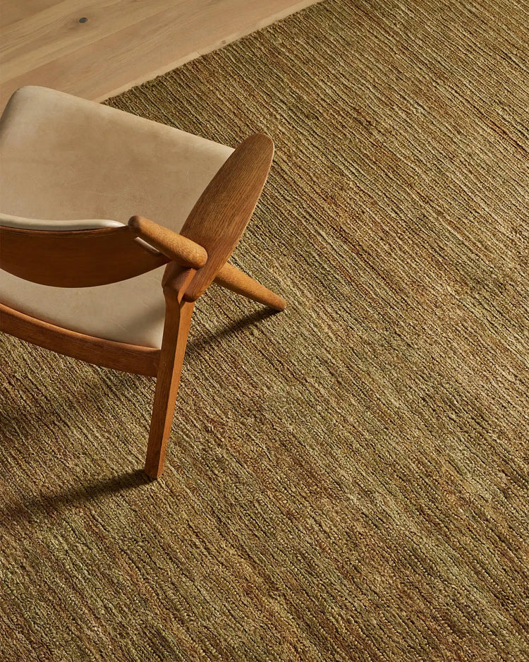 Mid-century wooden chair with cream upholstery on textured Bastian oakmoss jute rug, warm neutral palette with natural light casting shadows across woven floor covering
