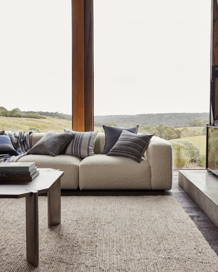 Neutral minimalist living room with cream boucle sofa, striped linen cushions, and woven area rug facing floor-to-ceiling windows overlooking rolling green hills and landscape views