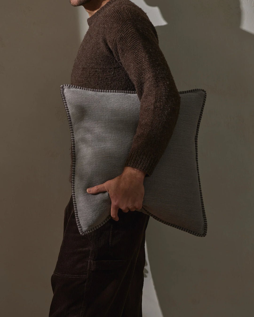 Man holding a grey linen cushion with whipstitched edge detail, wearing a dark brown knit sweater against neutral backdrop