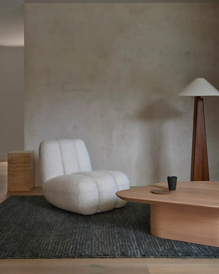 Minimalist lounge setting with charcoal jute rug, cream sculptural chair, and natural wood furniture against textured neutral walls