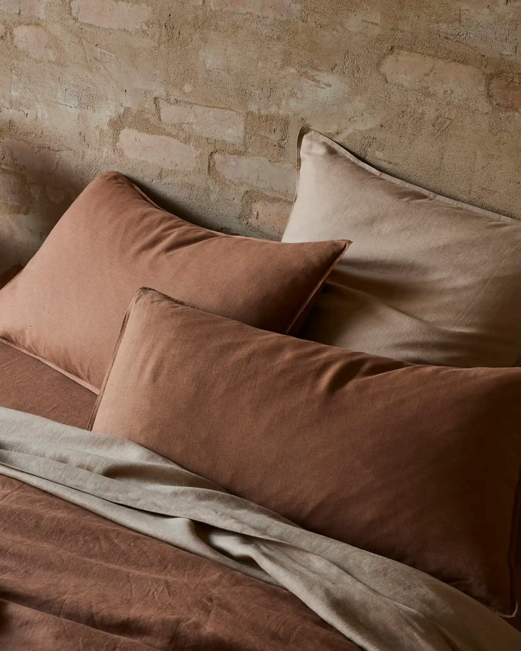 Neutral linen bedding styled on textured concrete wall, warm biscuit and cream tones layered with European linen pillows, showcasing soft, relaxed bedding texture