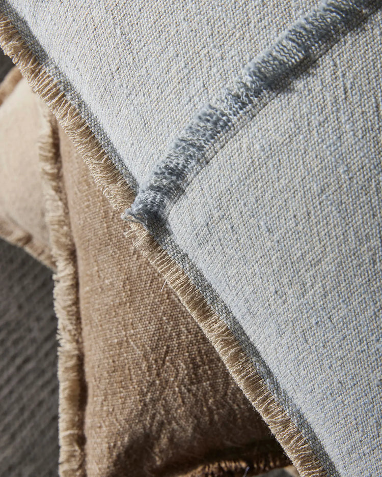 Close-up of layered cushions in neutral tones—soft grey-blue and warm taupe—with textured linen and cotton weave, fringe detailing, and natural sunlight highlighting the fabric composition