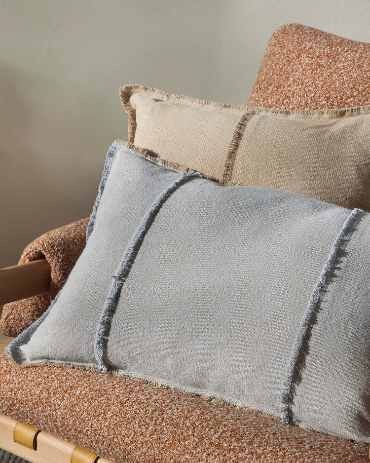 Warm terracotta armchair styled with layered cushions in soft blue-grey and cream linen, showcasing textured woven upholstery and fringed edge details in a neutral, earthy aesthetic