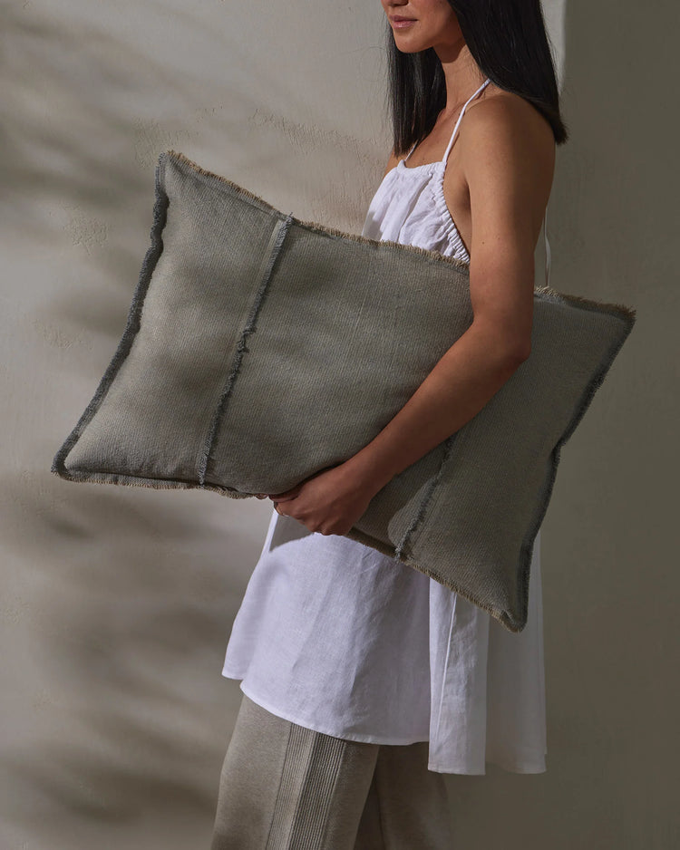 Sage linen cushions with raw frayed edges held against white linen, neutral minimalist styling with textured natural fibres and soft, earthy tones