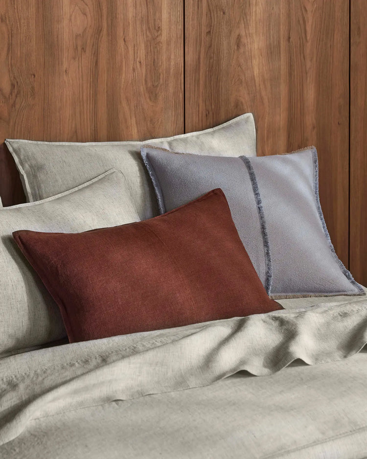 Warm timber bed styled with natural linen bedding, deep rosewood and soft haze cushions layered for an earthy, textured aesthetic with neutral pinstripe pillowcases