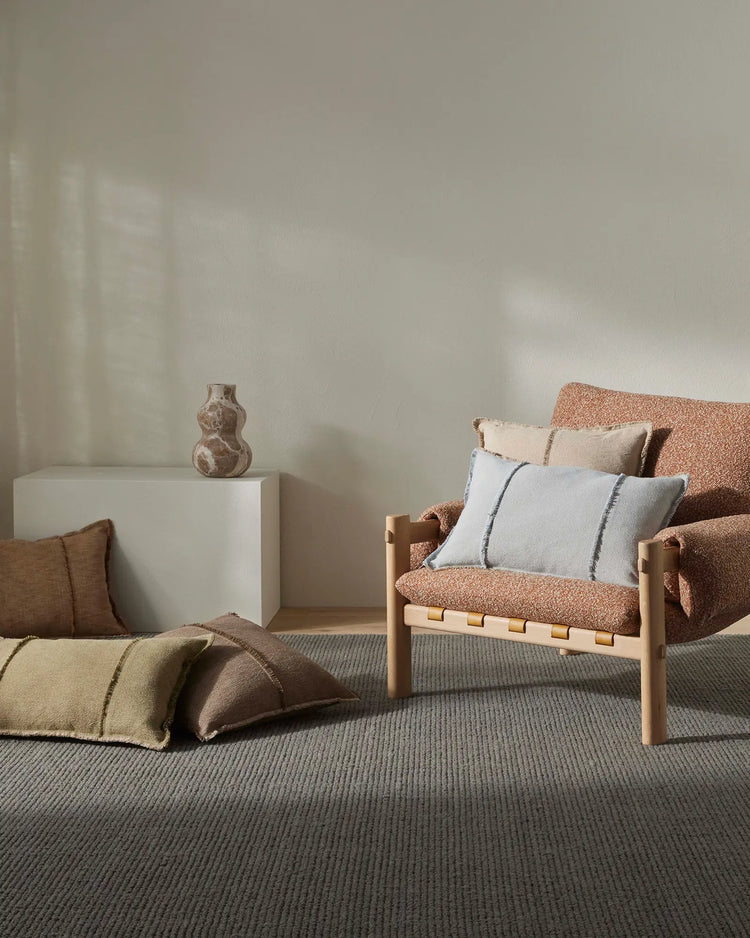 Neutral minimalist bedroom scene with terracotta and taupe textiles, natural wood furniture, striped grey carpet, and Adela cushions in soft cream and linen tones layered on woven armchair