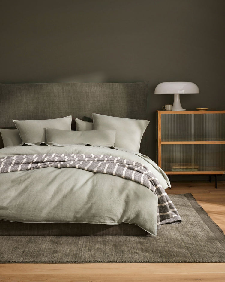 Earthy bedroom with sage linen bedding layered with a taupe check throw, grey upholstered headboard, mid-century timber nightstand, and natural wood flooring