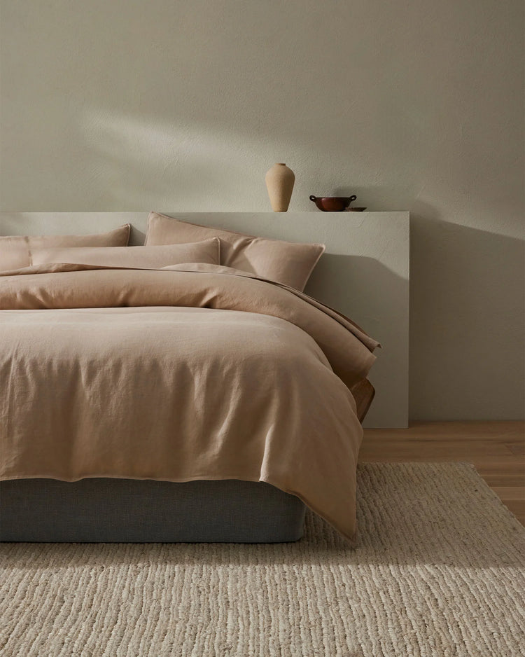 Minimalist bedroom with Ravello fitted sheet in warm shell tone, paired with natural linen textures and neutral palette. Cream bedding styled on charcoal base with woven jute rug and calming muted wall backdrop.