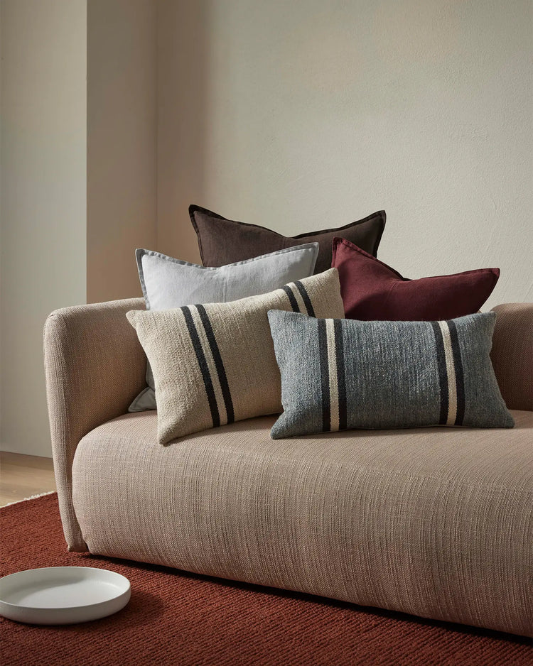 Neutral linen sofa layered with striped wool cushions in grey and cream, burgundy and charcoal accents, warm minimalist interior styling