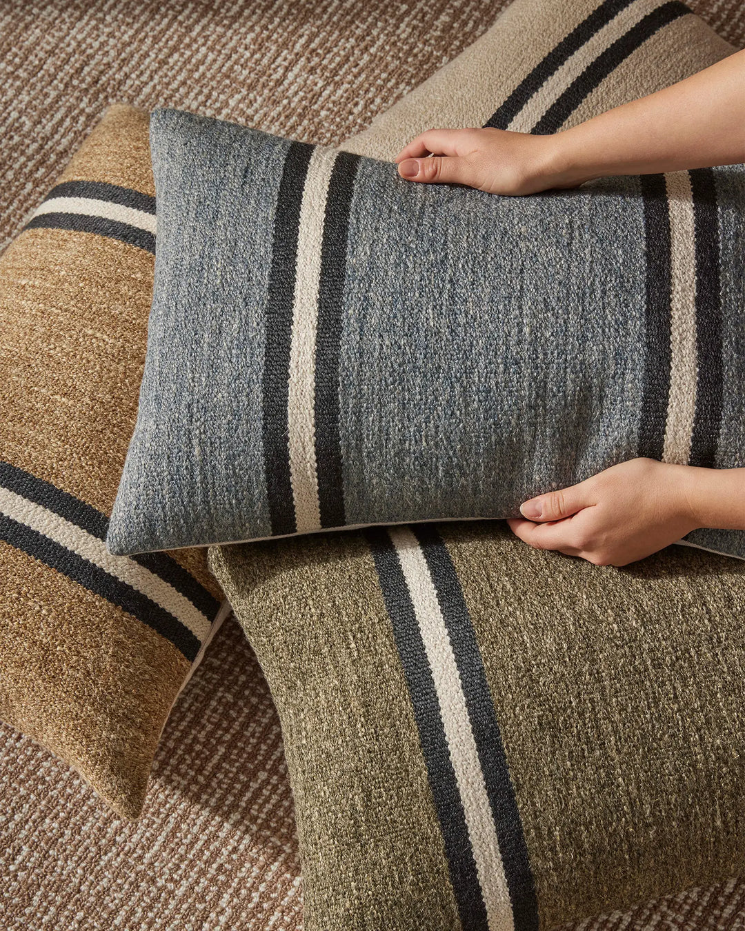 Neutral striped cushions in grey and natural tones layered on earth-toned textured rugs, showcasing woven wool and linen textures in a tactile, minimalist home setting