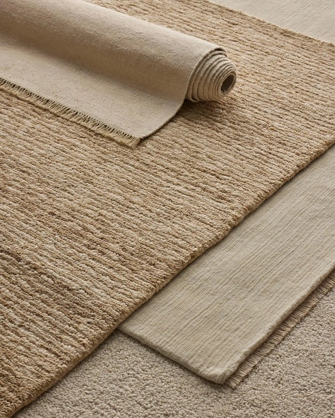 Neutral layered rugs in cream and beige tones showcase textural contrast—a rolled canvas rug alongside woven jute and wool blend options in warm earthy hues