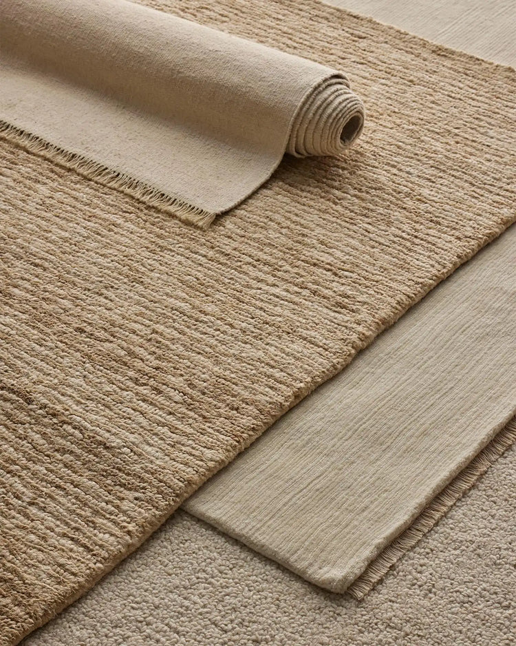 Neutral layered rugs in cream and beige tones showcase textural contrast—a rolled canvas rug alongside woven jute and wool blend options in warm earthy hues