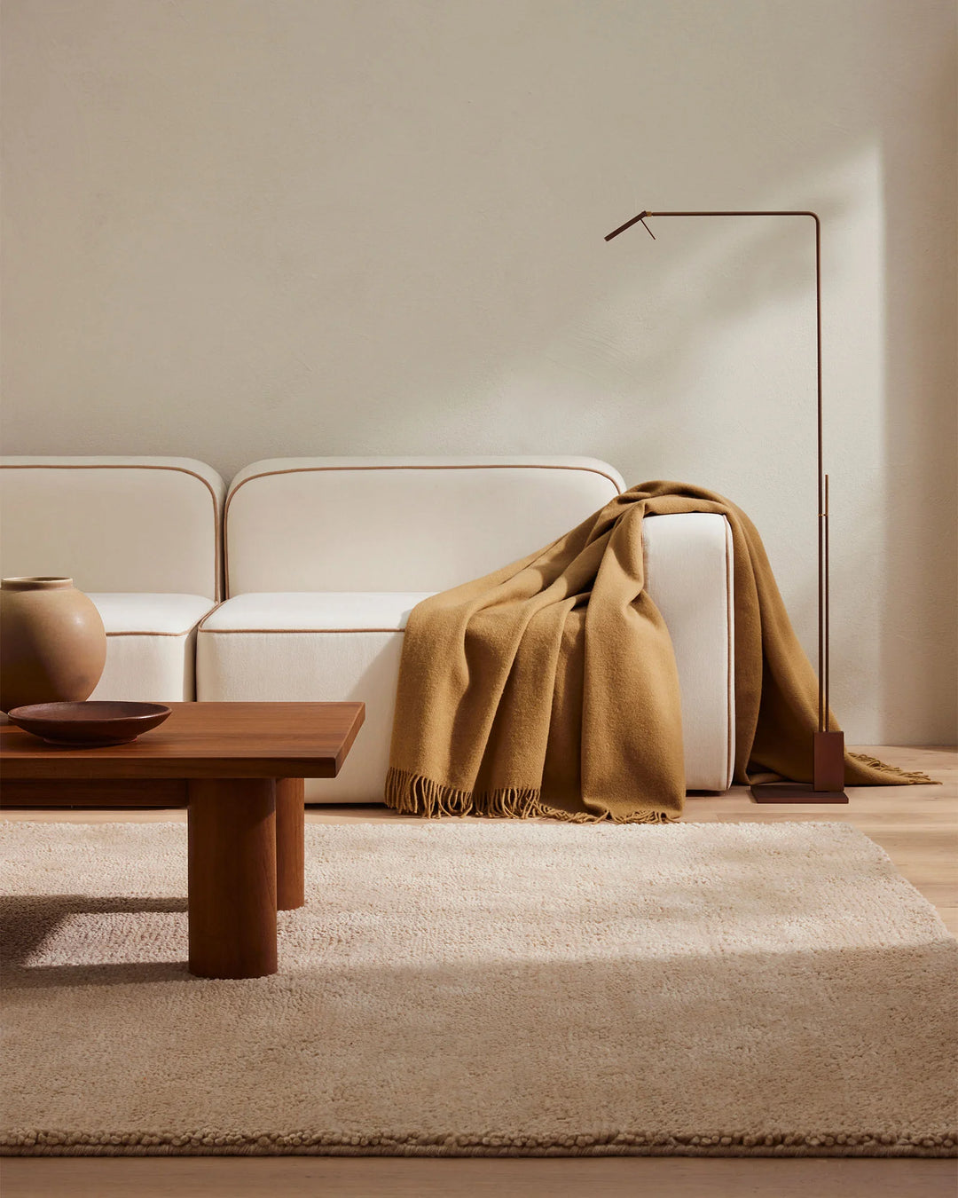 Warm minimalist living room with neutral cream sofa, golden mustard wool throw, natural wood furniture, and layered terracotta rugs in earthy tones