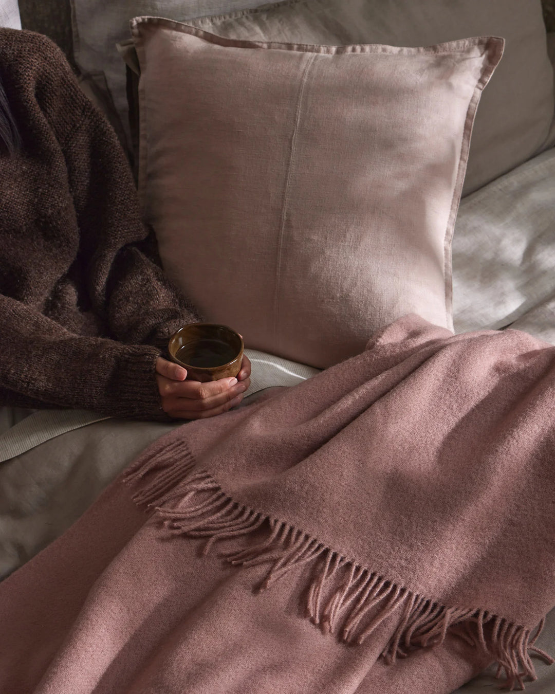 Mauve and blush toned lifestyle scene with Nevis throw featuring fringe detail and Como cushion on neutral bedding, soft natural light