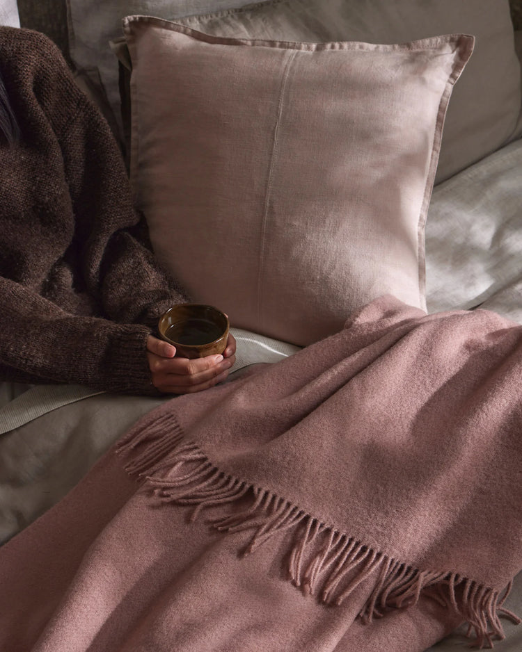 Mauve and blush toned lifestyle scene with Nevis throw featuring fringe detail and Como cushion on neutral bedding, soft natural light