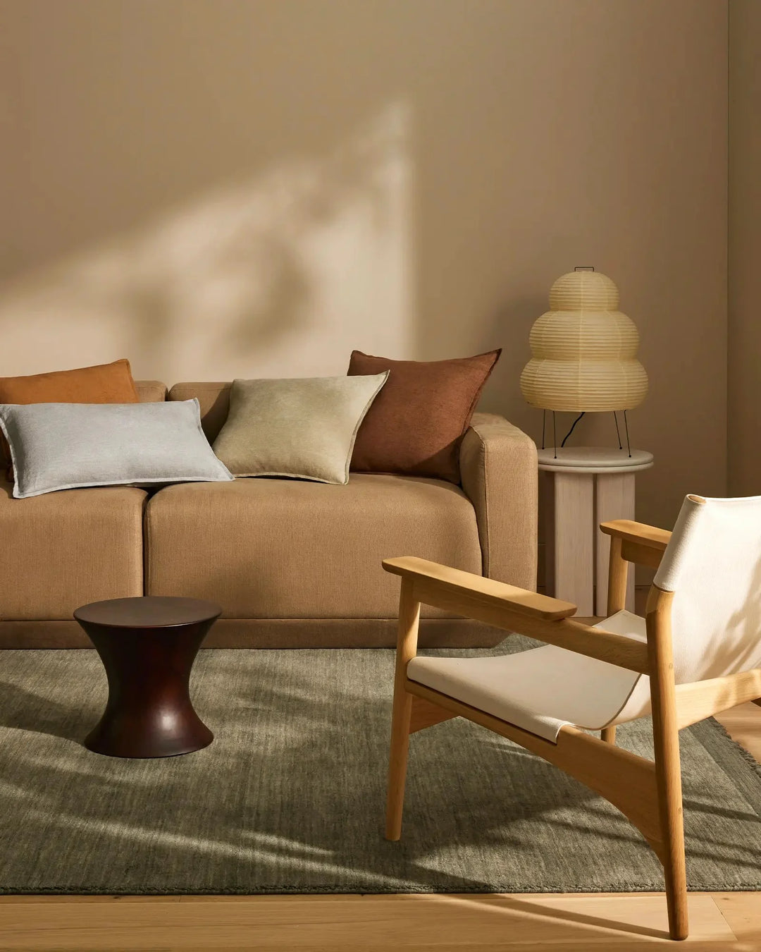 Warm earthy living room with caramel sofa and Fiore cushions in cream, sage and rust tones; natural light, timber chair, neutral rug and paper pendant lamp create minimalist, organic aesthetic.