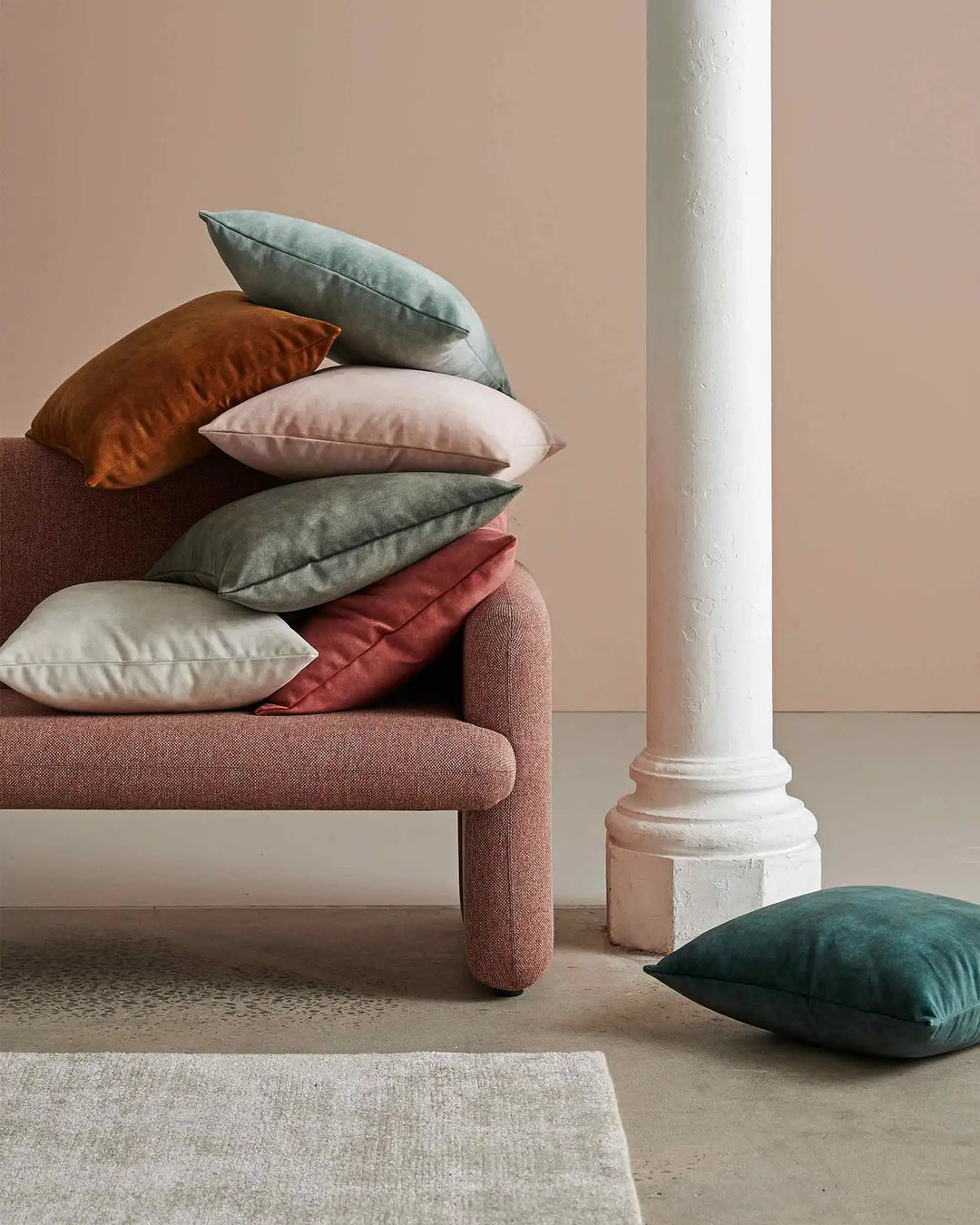 Earthy toned living room styling with stacked Ava cushions in rust, sage, cream and terracotta on a mauve upholstered bench, beside a white architectural column and neutral textured rug