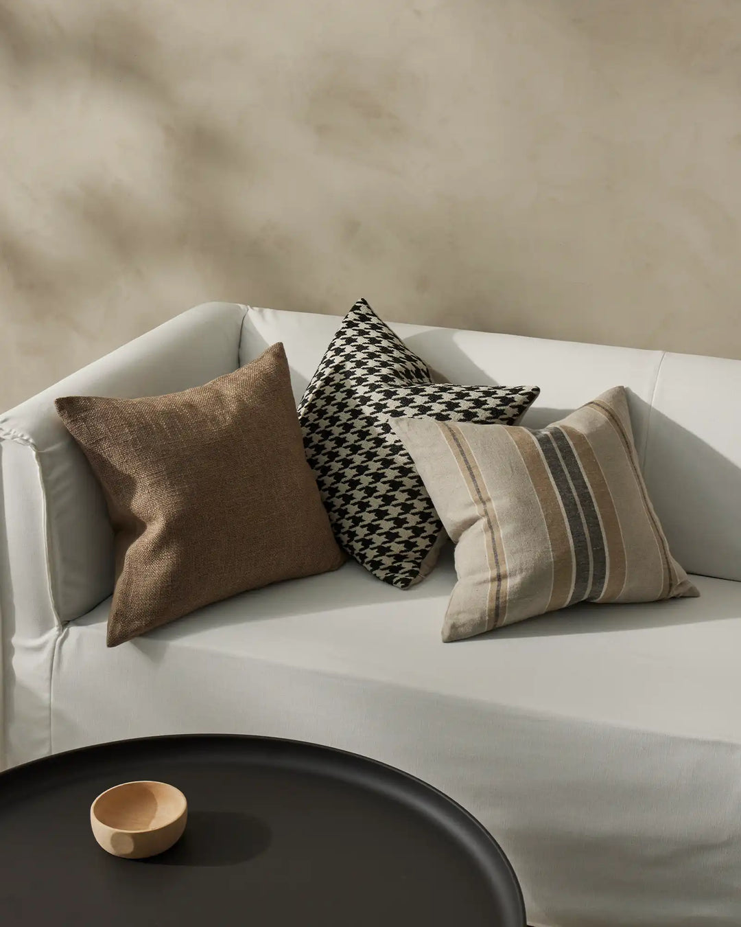 Neutral modern bedroom with white curved sofa styled with three cushions: earthy textured Domenica, monochrome houndstooth Giovanni, and striped Franco in warm naturals and greys