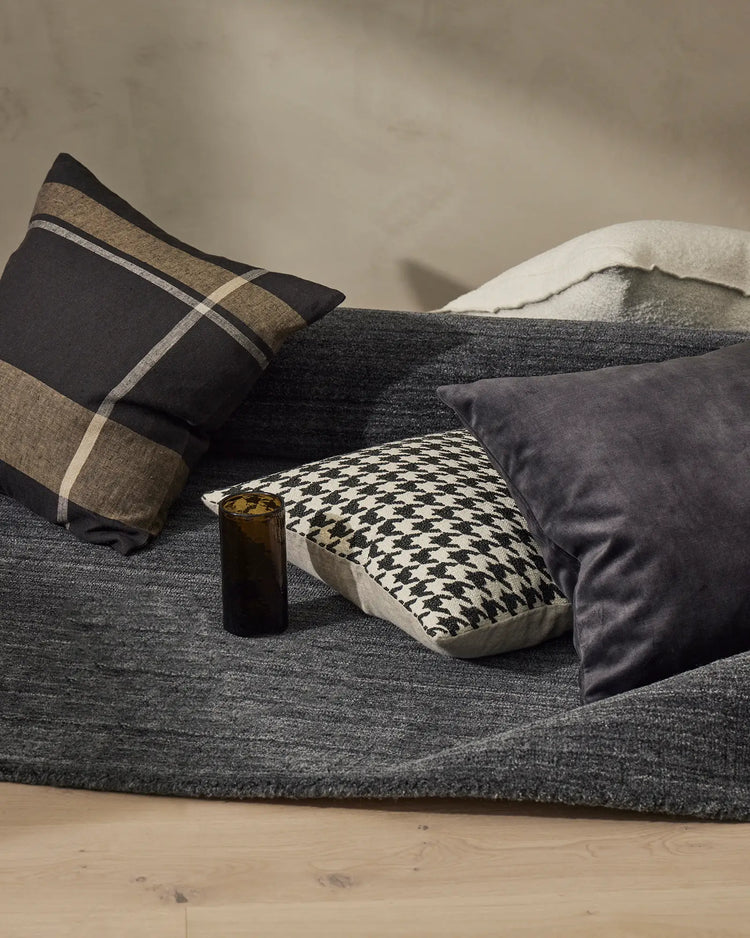 Neutral bedroom styling with dark charcoal sofa, geometric patterned cushion, striped throw pillow in black and tan, cream knit blanket, and moody grey palette with natural wood flooring