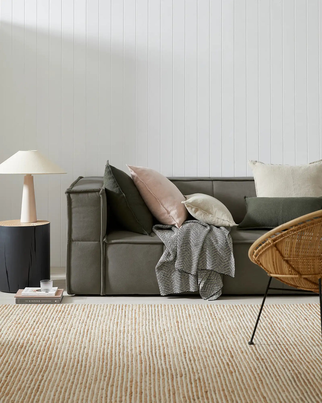 Neutral living room with olive-green sofa styled with cream and mauve pillows, black and white herringbone throw, natural jute rug, rattan chair, and minimalist furnishings against white shiplap wall