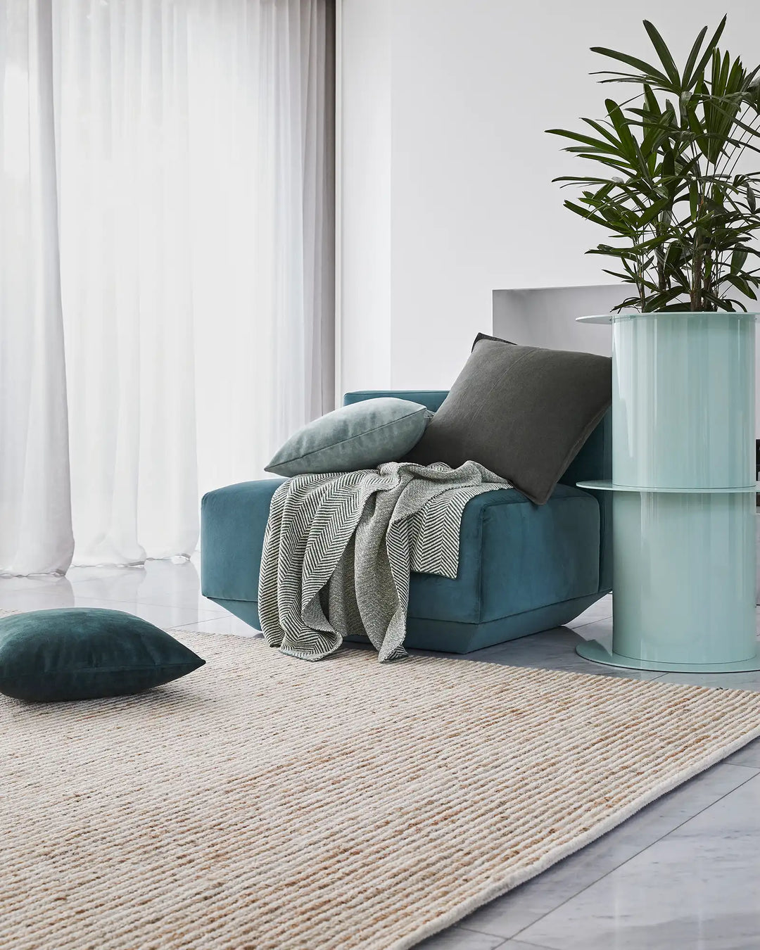 Scandinavian living room with teal upholstered chair, herringbone throw, grey cushions and Lisbon jute rug in natural tones, brightened by sheer white curtains and potted green plant