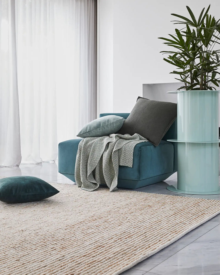 Scandinavian living room with teal upholstered chair, herringbone throw, grey cushions and Lisbon jute rug in natural tones, brightened by sheer white curtains and potted green plant