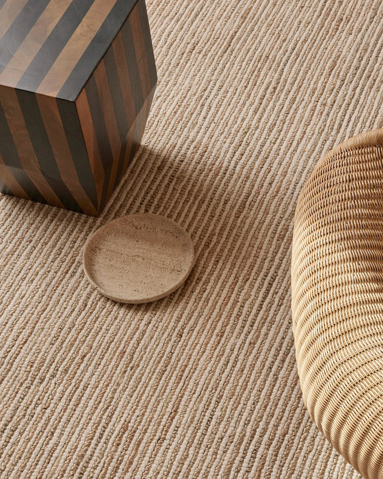 Neutral jute rug with bold striped wooden stool and woven rattan chair leg, earthy palette with textured surfaces