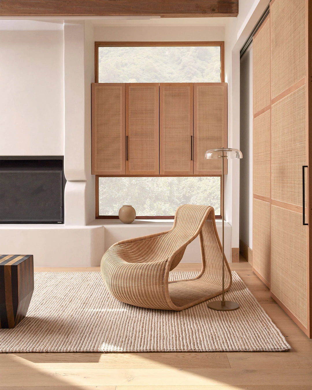 Neutral minimalist bedroom with warm timber and rattan textures, featuring Lisbon Seasalt jute rug anchoring sculptural woven chair beneath tall windows