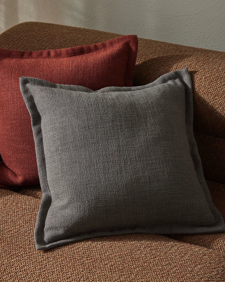 Charcoal and burgundy cushions layered on natural textured lounge chair, warm earth tones and soft linen fabrics create cosy minimalist living room styling