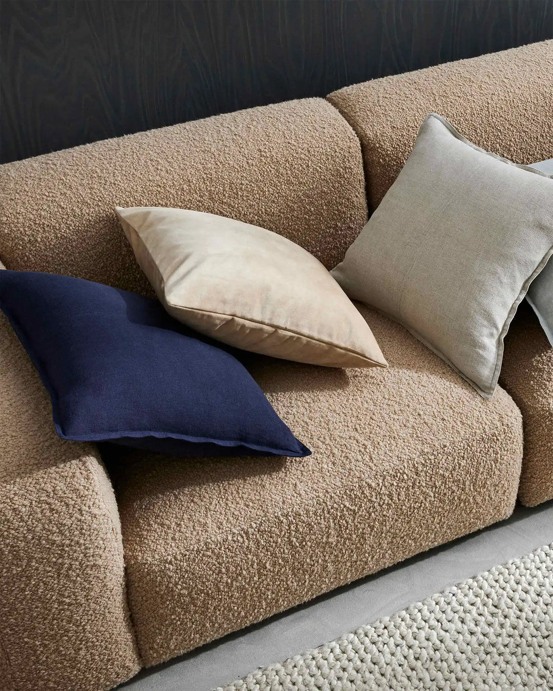 Neutral linen and textured cushions in cream, navy and beige arranged on a nougat-toned sofa with dark timber wall backdrop and cream area rug