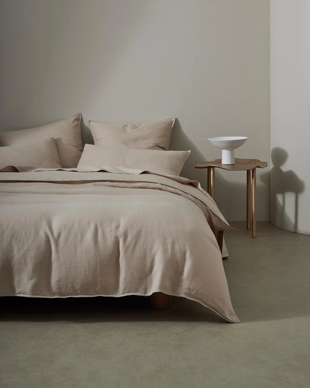 Minimalist bedroom with Ravello flat sheet and pillowcases in soft shell linen, layered neutral tones and relaxed textures against pale walls, mid-century timber side table.