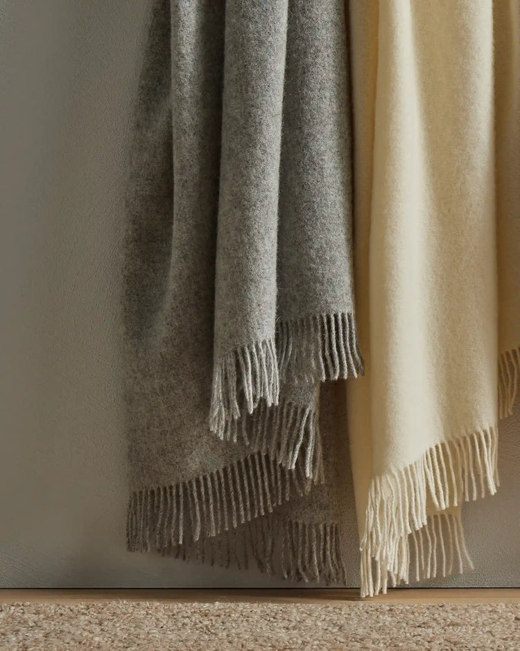 Nevis throws in ash and snow draped against neutral wall, showcasing soft wool textures and hand-fringed edges in warm greyscale and cream tones