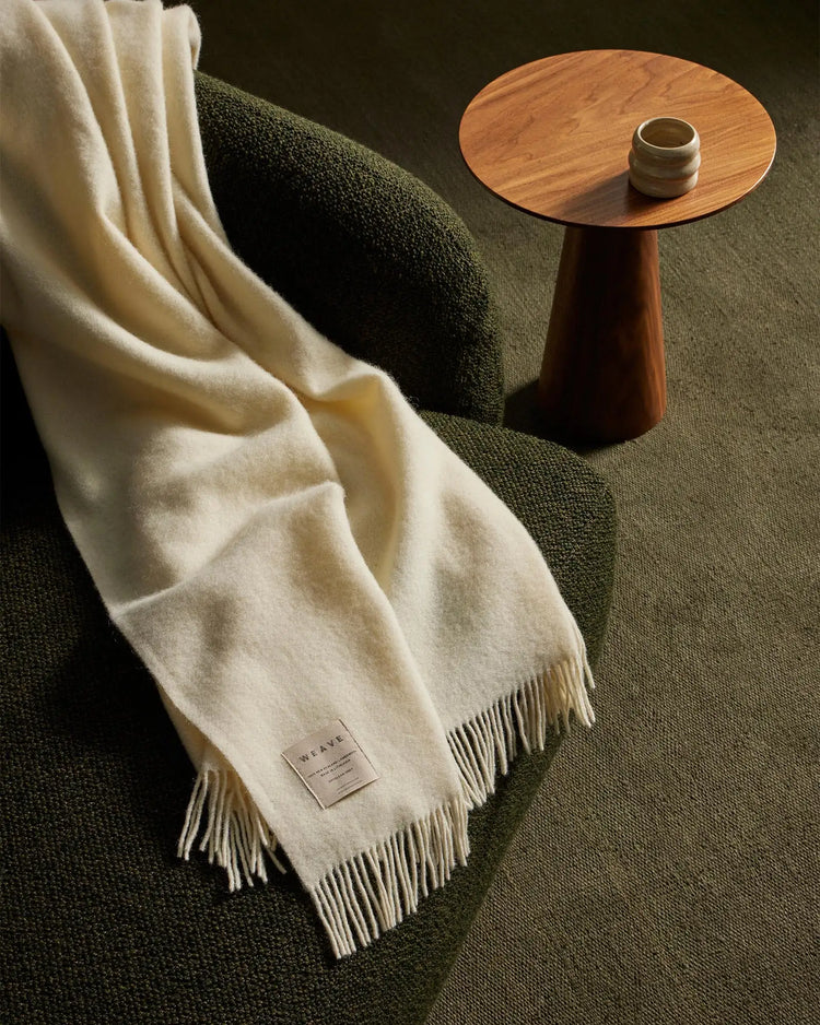 Cream wool throw draped over sage green upholstered chair, minimalist wood side table, earthy natural palette with soft textures and neutral tones
