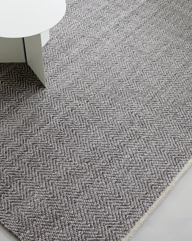 Matterhorn wool-cotton blend rug in charcoal, featuring geometric chevron weave texture, anchoring a minimalist interior with soft greige tones and modern furnishings